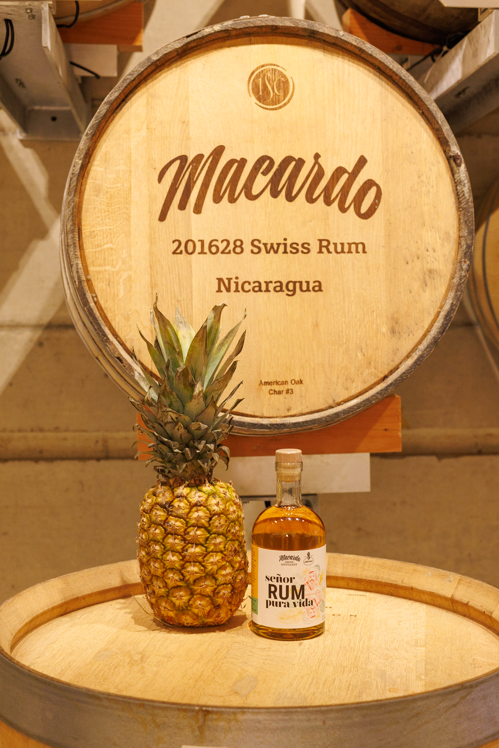 Ein Rum-Flasche "Señor Rum - Pura Vida" und eine Ananas stehen auf einem hellen Holzfass. Im Hintergrund ist ein zweites Fass mit der Aufschrift "Macardo Swiss Rum Nicaragua" zu sehen.